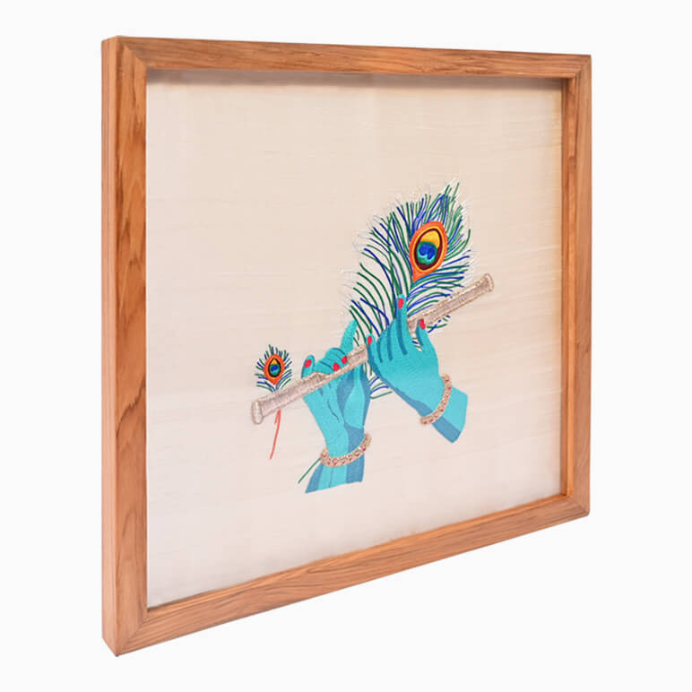 Left angled view of a Krishna flute artwork, hand embroidered in blue and gold over a cream fabric background, seen framed in wood