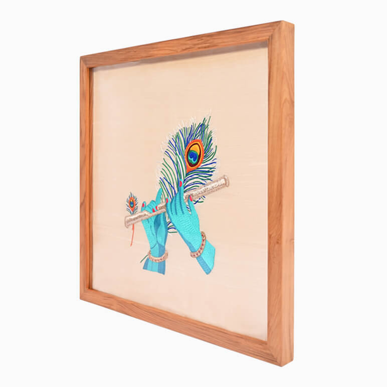 Right angled view of a Krishna flute artwork, hand embroidered in blue and gold over a cream fabric background, seen framed in wood