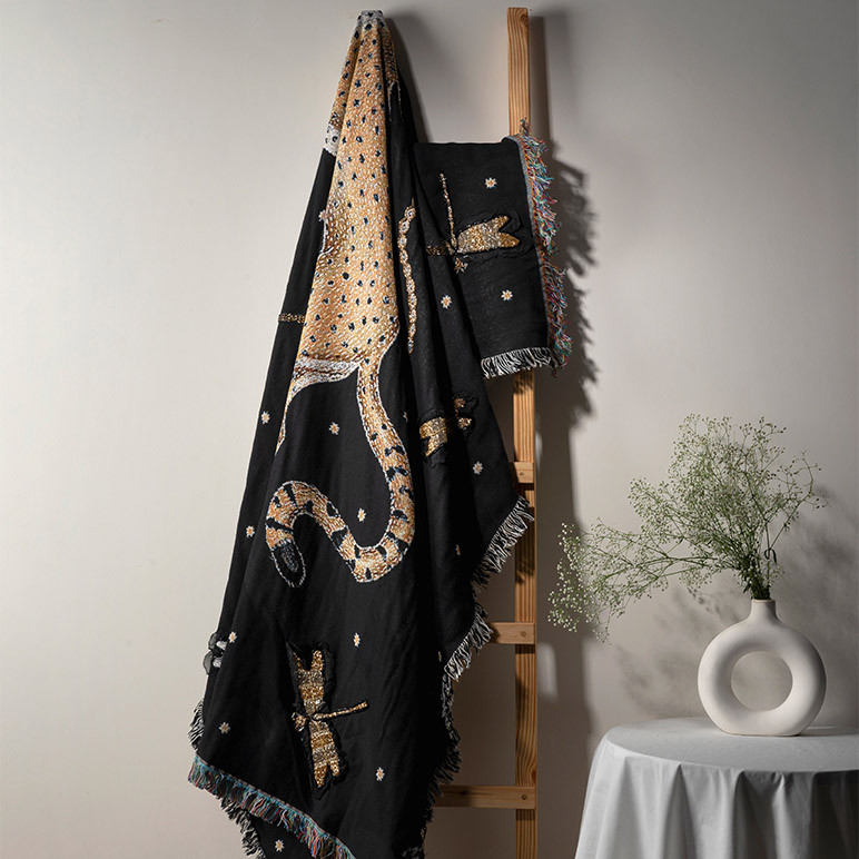 Black colored woven throw blanket artistically laid on a ladder by a white table with a vase 