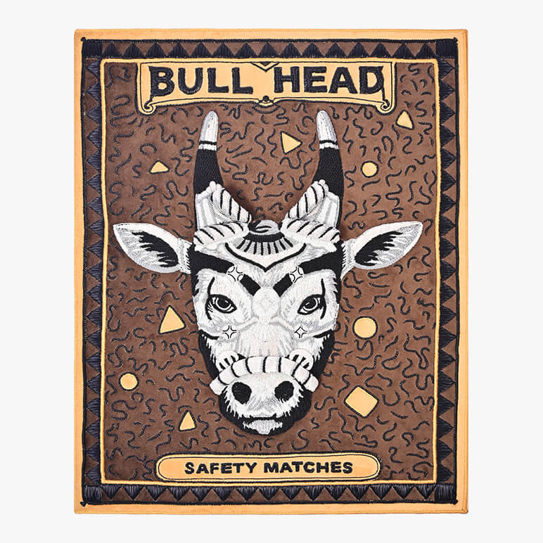 framed 3d textile wall art showcasing an enlarged vintage matchbox crafted using metallic embroidery to form a sculpted bull's head decorated with ornaments, seen over an earthy brown suede fabric. Has the titles 