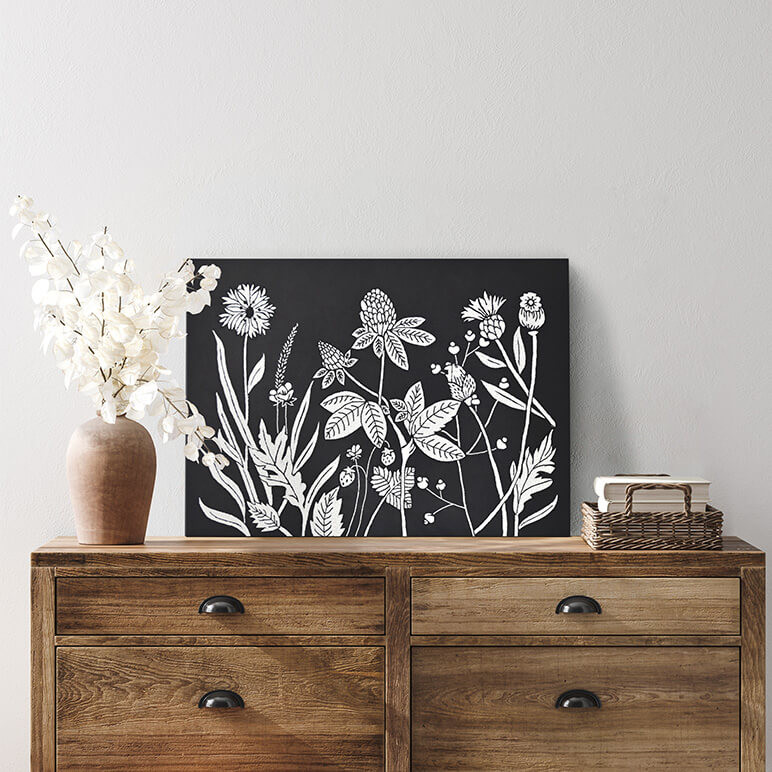 Mood shot of a minimalist wall art with wildflowers, in black and white colors, seen leaning against a wall over a wooden console table