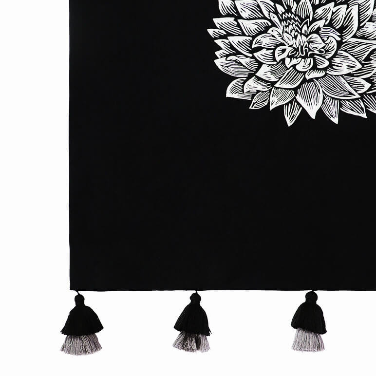 corner view of a monochrome wall art featuring a black tapestry with tassels and a large white embroidered dahlia flower at the center