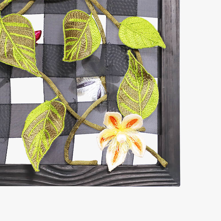 view of a corner of a framed textile wall artwork inspired by nature, featuring a bunch of green embroidered leaves and a peach colored Clematis flower