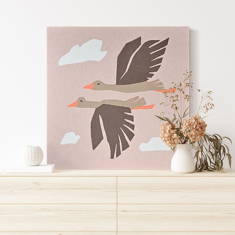 Staged view of a nature inspired textile wall art piece featuring two flying birds amidst a pink sky, seen displayed over a console table