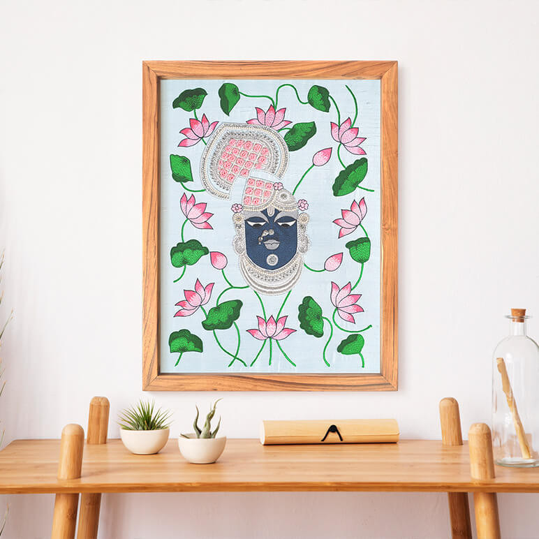 Staged view of a wooden framed Shrinathji textile artwork, seen displayed over a white wall behind a wooden table