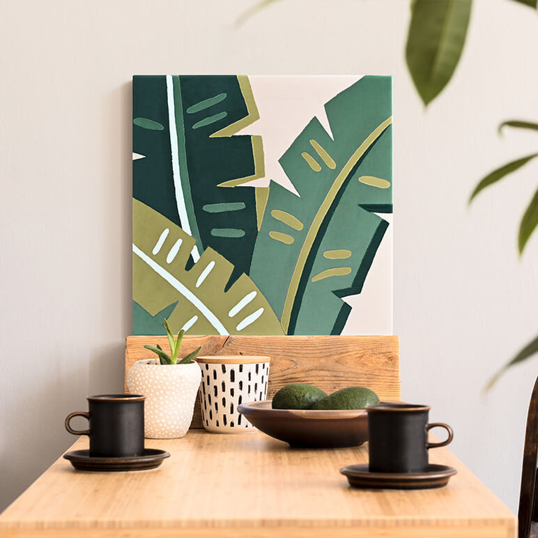 Staged view of a tropical wall art featuring green toned banana leaves in patchworked form, canvas framed and displayed by a wooden dining table