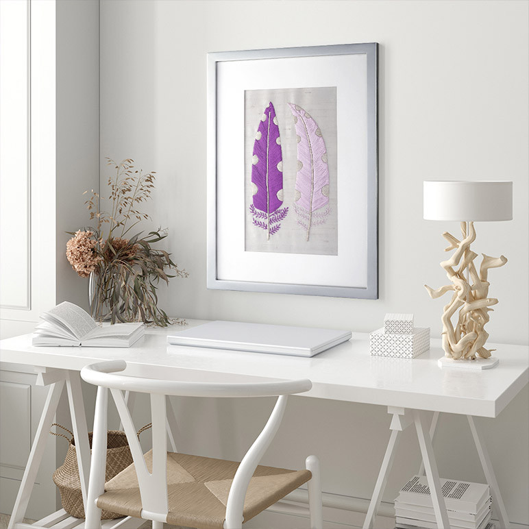 framed textile art featuring two purple feathers in a white room over a white table
