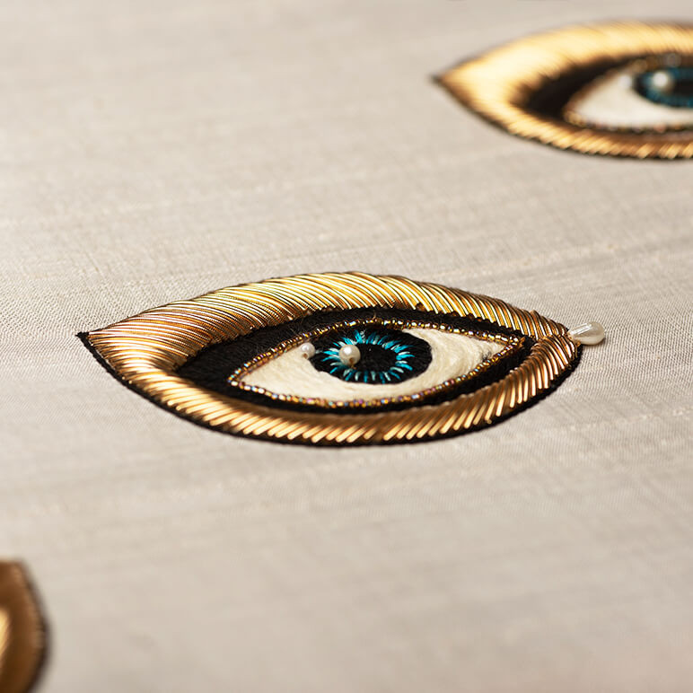 closeup embroidery detail showing a gold metallic embroidered blue eye over an off-white silk background